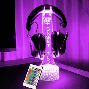 May include: A white LED light stand with a custom gamer tag, "GAMEKTAG", and a clear acrylic headset holder. The stand has a white, textured base and is illuminated in a vibrant purple color. A remote control with color options is included.