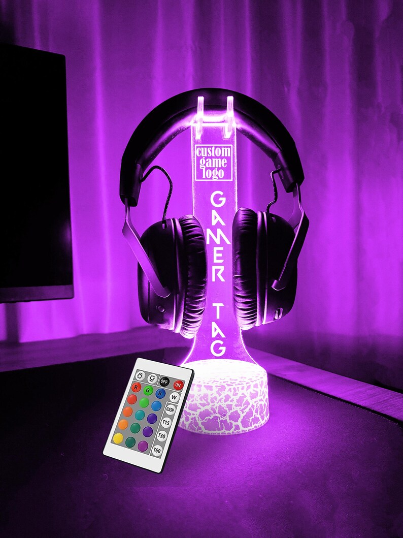 May include: A white LED light stand with a custom gamer tag, "GAMEKTAG", and a clear acrylic headset holder. The stand has a white, textured base and is illuminated in a vibrant purple color. A remote control with color options is included.