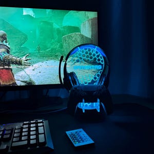 May include: A blue and black gaming headset stand with a blue light and the word "GAMERTAG" on the front. The stand is on a black surface with a keyboard and a remote control.