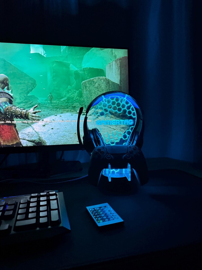 May include: A blue and black gaming headset stand with a blue light and the word "GAMERTAG" on the front. The stand is on a black surface with a keyboard and a remote control.