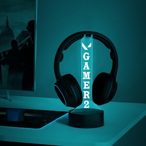 May include: A black gaming headset sits on a glowing LED stand. The stand displays the word "GAMER2" in white. The setup is on a white desk, with a computer monitor in the background. The lighting has a cool blue hue.