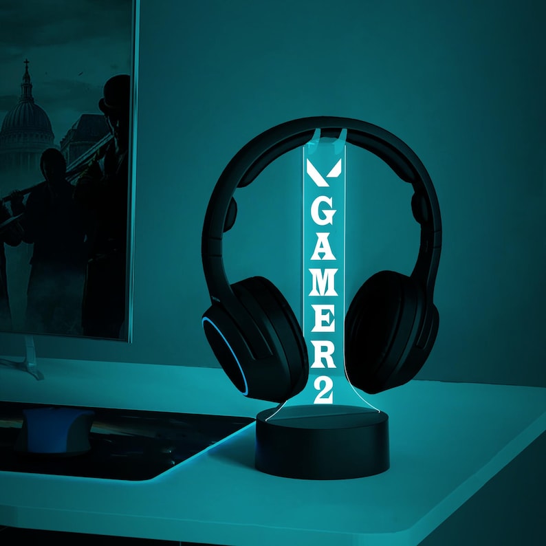 May include: A black gaming headset sits on a glowing LED stand. The stand displays the word "GAMER2" in white. The setup is on a white desk, with a computer monitor in the background. The lighting has a cool blue hue.