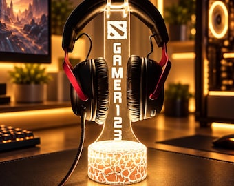 May include: A clear acrylic headphone stand with a glowing base featuring a cracked texture pattern. The vertical support displays the text GAMER123 and a stylized logo. Black and red over-ear headphones are resting on the top of the stand.