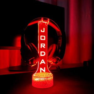 May include: A red LED light with a custom nameplate that reads "JORDAN" and a headphone stand. The light has a textured base.