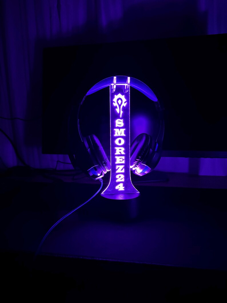May include: A purple LED light-up headset stand with the name "SMOREZ24" and a symbol on it. The stand is holding a black headset.