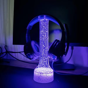 Op de afbeelding: Een paarse LED-lamp met een printplaatontwerp, die een zwarte koptelefoon vasthoudt. De lamp heeft een getextureerde basis.