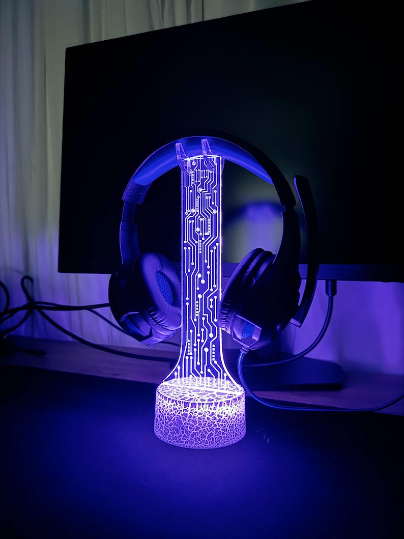 Op de afbeelding: Een paarse LED-lamp met een printplaatontwerp, die een zwarte koptelefoon vasthoudt. De lamp heeft een getextureerde basis.