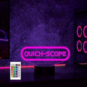 May include: A pink neon sign with the text "QUICK-SCOPE" in a retro video game style. The sign is on a black base and has a remote control with multiple colored lights.
