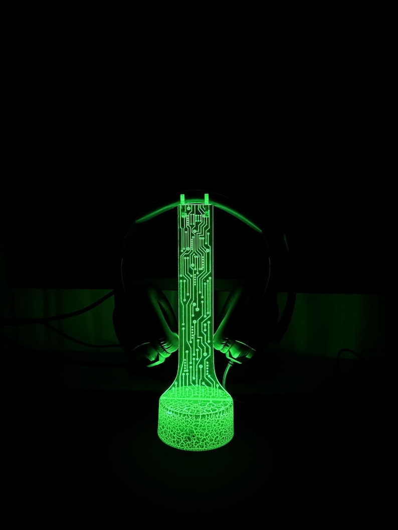 Puede incluir: Una l&aacute;mpara LED verde con un dise&ntilde;o de placa de circuito, sentada sobre una superficie negra. La l&aacute;mpara est&aacute; rodeada de auriculares negros.