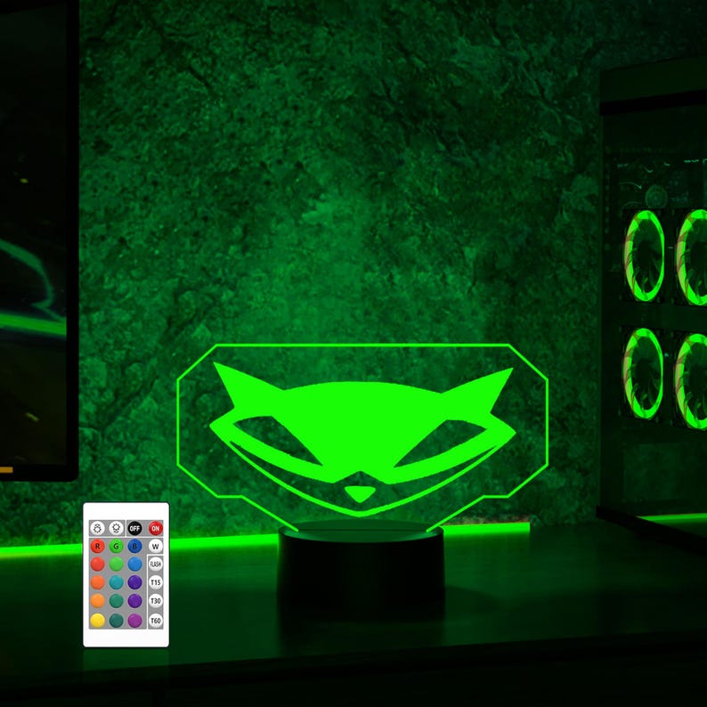 May include: A green raccoon face logo illuminated by a 3D LED lamp with a remote control. The lamp is on a black base and has a green glow.