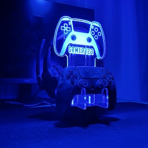 Op de afbeelding: Een blauw verlichte gamecontrollerstandaard met de tekst "GAMER123" op het controller-vormige licht. Een camouflage-patroon controller rust in de standaard, met een headset erachter. De standaard staat op een donkere ondergrond.