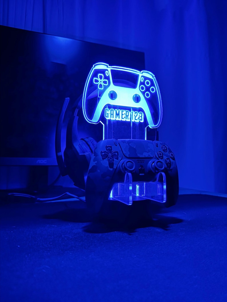 Op de afbeelding: Een blauw verlichte gamecontrollerstandaard met de tekst "GAMER123" op het controller-vormige licht. Een camouflage-patroon controller rust in de standaard, met een headset erachter. De standaard staat op een donkere ondergrond.