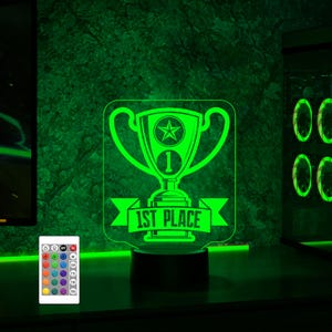 Puede incluir: Una luz nocturna LED verde en forma de trofeo con una estrella y el texto "1st Place". La luz est&aacute; sobre una base negra y tiene un mando a distancia con m&uacute;ltiples opciones de color.