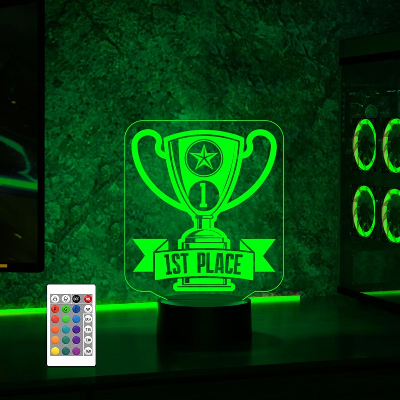 Puede incluir: Una luz nocturna LED verde en forma de trofeo con una estrella y el texto "1st Place". La luz est&aacute; sobre una base negra y tiene un mando a distancia con m&uacute;ltiples opciones de color.