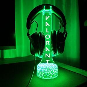 May include: A green LED light-up stand with a cracked glass base, holding a pair of black headphones. The stand has the word "VALORANT" written on it in white.