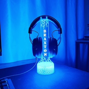 May include: A black headset rests on a blue-lit stand. The stand features the name "BRAXTON" in white lettering and a lightning bolt design. The base of the stand has a cracked glass effect, and the background is a solid blue.