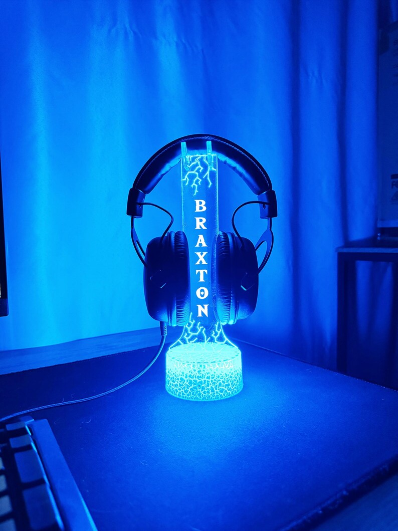 May include: A black headset rests on a blue-lit stand. The stand features the name "BRAXTON" in white lettering and a lightning bolt design. The base of the stand has a cracked glass effect, and the background is a solid blue.