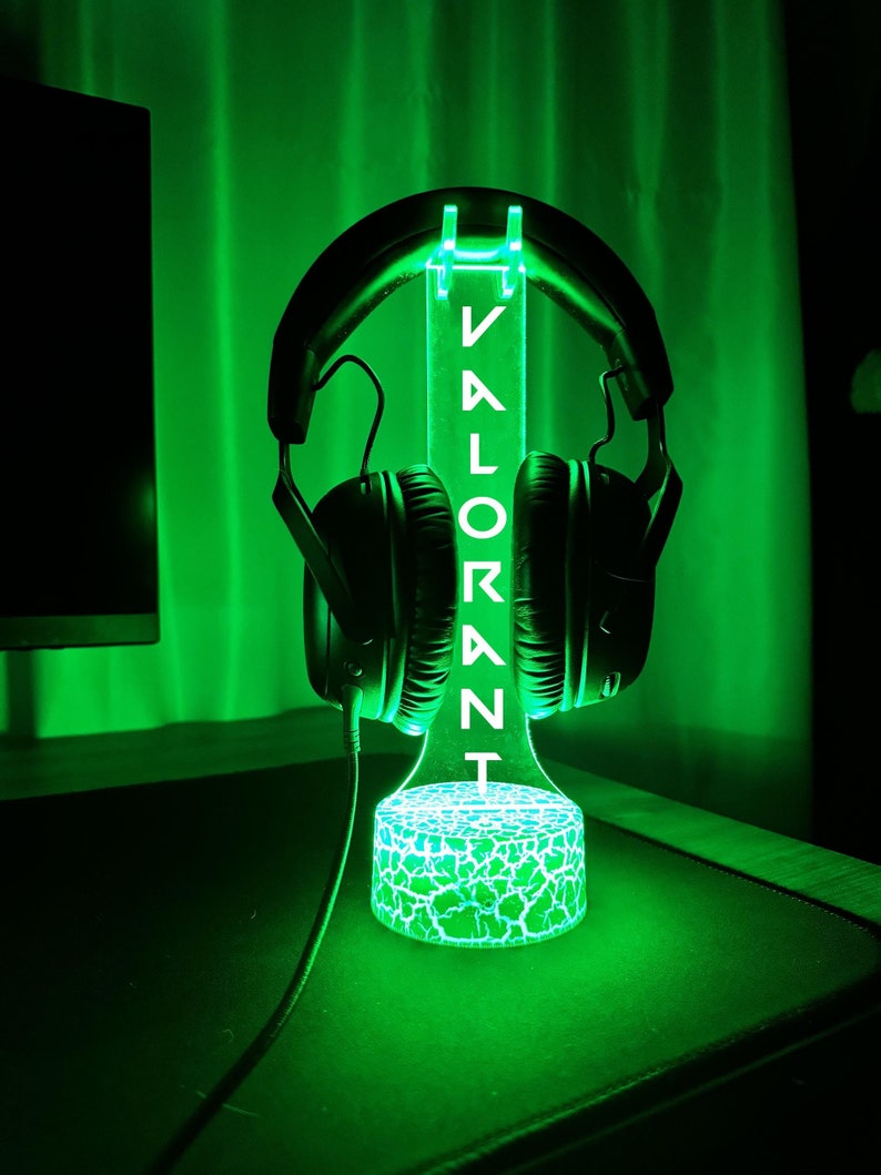 May include: A green-lit headphone stand with the word "VALORANT" vertically displayed. The stand holds a pair of black headphones. The base of the stand has a cracked, glowing design. The background is a solid green.