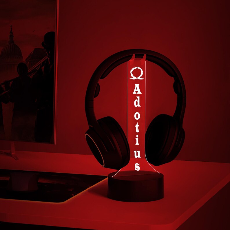 May include: A black headset resting on a custom-made LED stand. The stand is illuminated with a bright white light, displaying the name "Adotius" vertically. The background is a dark red, creating a dramatic effect.