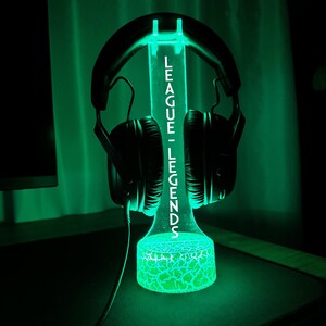 K&ouml;nnte beinhalten: Ein gr&uuml;n leuchtender Kopfh&ouml;rerst&auml;nder mit dem wei&szlig;en Schriftzug "LEAGUE - LEGENDS". Ein schwarzer Kopfh&ouml;rer ist darauf platziert. Der Sockel hat ein Rissglas-Design. Der St&auml;nder steht auf einer dunklen Oberfl&auml;che.