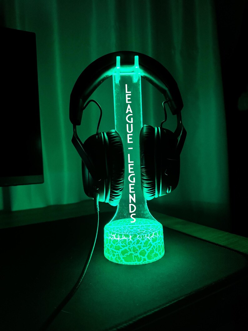 K&ouml;nnte beinhalten: Ein gr&uuml;n leuchtender Kopfh&ouml;rerst&auml;nder mit dem wei&szlig;en Schriftzug "LEAGUE - LEGENDS". Ein schwarzer Kopfh&ouml;rer ist darauf platziert. Der Sockel hat ein Rissglas-Design. Der St&auml;nder steht auf einer dunklen Oberfl&auml;che.