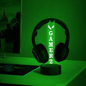May include: A black headset sits on a green illuminated stand. The stand displays the word "GAMER2" in white. The setup is on a green surface, creating a gaming aesthetic. The lighting is green.