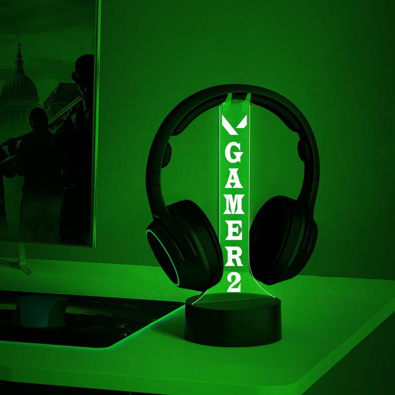 May include: A black headset sits on a green illuminated stand. The stand displays the word "GAMER2" in white. The setup is on a green surface, creating a gaming aesthetic. The lighting is green.