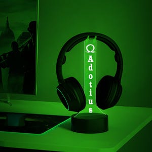 May include: A black headset rests on a glowing green stand. The stand has the word "Adotius" vertically displayed in white. The background is a dark green, with a computer monitor visible on the left side.