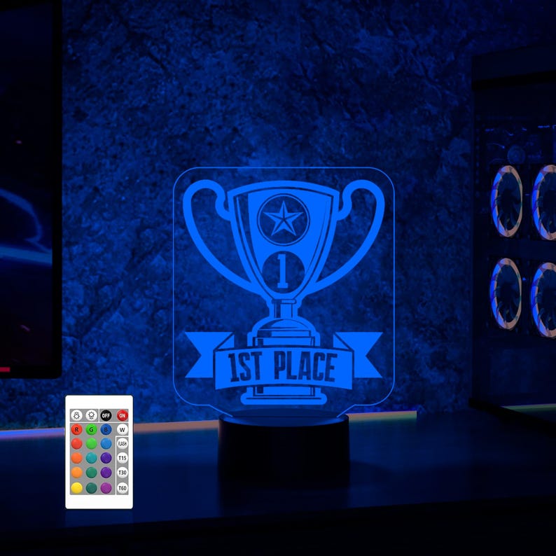 Puede incluir: Una luz nocturna de acr&iacute;lico azul con forma de trofeo con una estrella y el texto "1st Place". La luz est&aacute; sobre una base negra y tiene un mando a distancia con m&uacute;ltiples opciones de color.