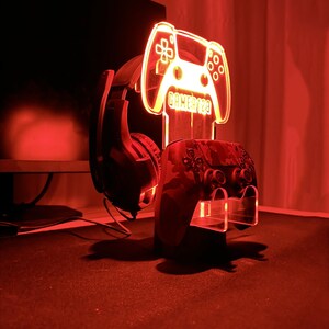 Op de afbeelding: Een rood verlichte gaming-opstelling met een controllerstandaard, een headset en een controller met camouflagepatroon. De standaard heeft de vorm van een gamecontroller met de tekst "GAMER123". De opstelling wordt verlicht door een rode gloed.