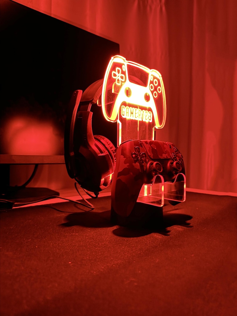 Op de afbeelding: Een rood verlichte gaming-opstelling met een controllerstandaard, een headset en een controller met camouflagepatroon. De standaard heeft de vorm van een gamecontroller met de tekst "GAMER123". De opstelling wordt verlicht door een rode gloed.
