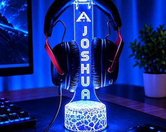 May include: A clear acrylic headphone stand with a glowing blue LED base featuring a cracked texture. The vertical support displays the name JOSHUA in white capital letters below a triangular logo. Black over-ear headphones rest on top.