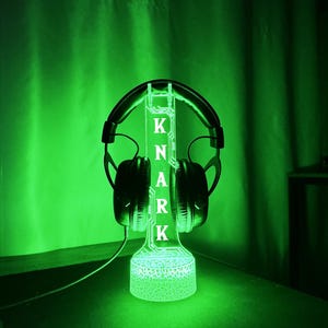 May include: A green-glowing headphone stand with the name "KNARK" illuminated in white. Black headphones rest on the stand. The stand has a textured base and a vertical support with the letters. The background is a dark green.
