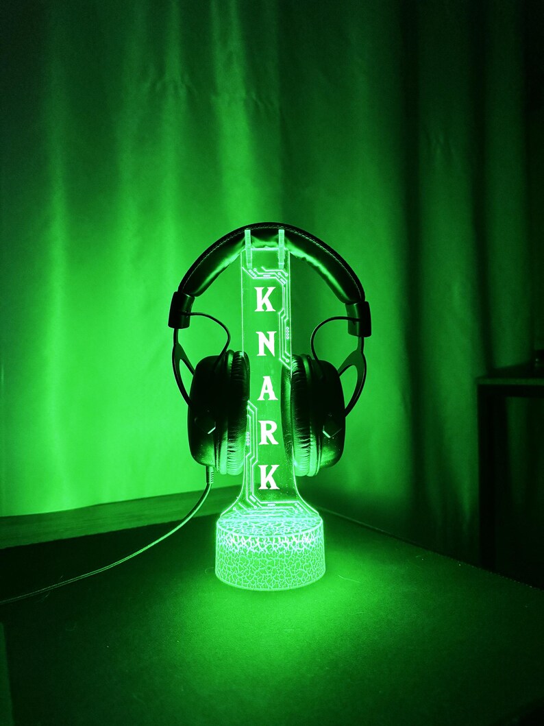 May include: A green-glowing headphone stand with the name "KNARK" illuminated in white. Black headphones rest on the stand. The stand has a textured base and a vertical support with the letters. The background is a dark green.