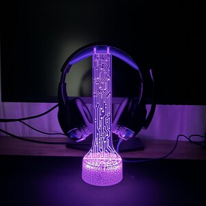 Puede incluir: Una luz LED p&uacute;rpura con un dise&ntilde;o de placa de circuito y una base texturizada. La luz est&aacute; dise&ntilde;ada para sostener auriculares.