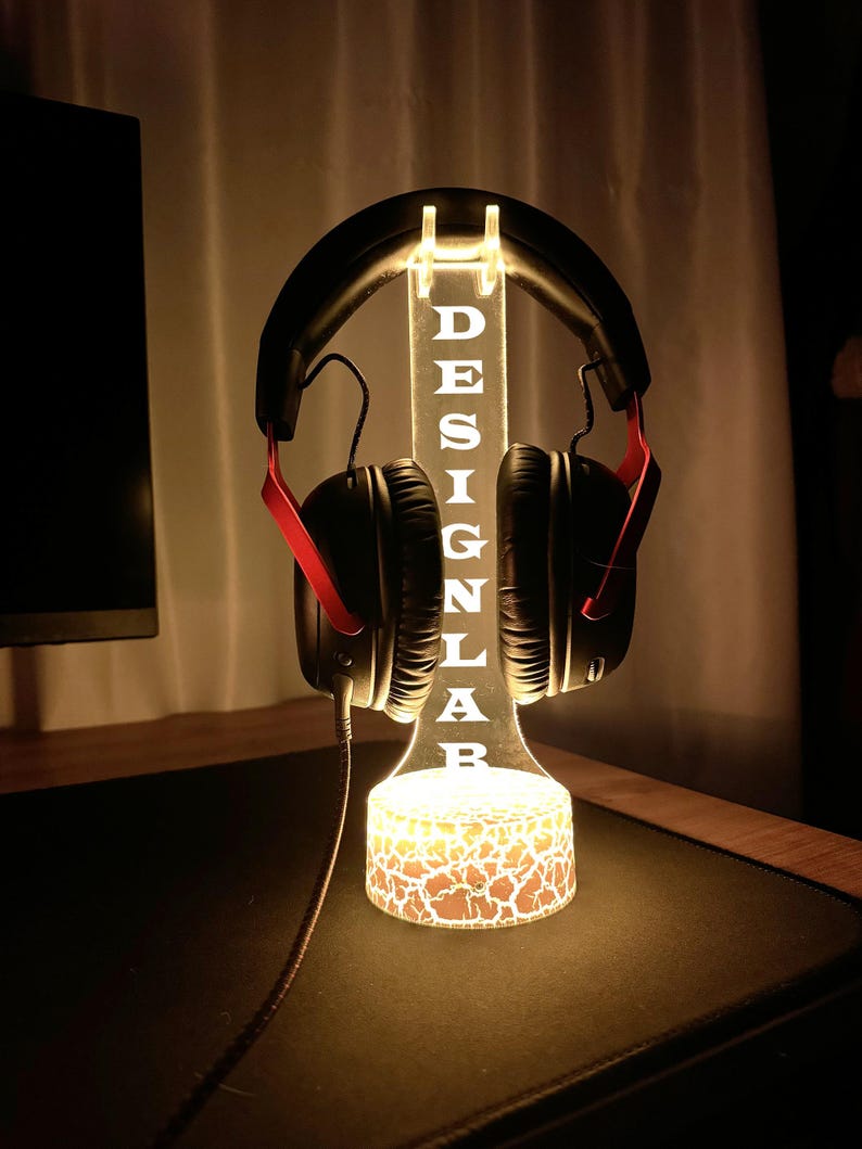 May include: A lighted headphone stand with the word "DESIGN LAB" etched into the acrylic. The stand is illuminated with a warm glow, and a pair of black headphones with red accents are displayed. The base has a cracked earth design.