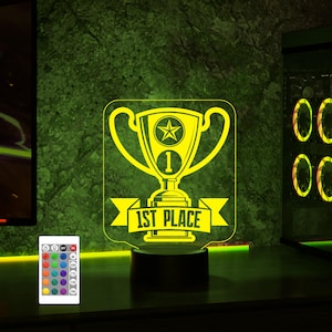Puede incluir: Una luz nocturna LED amarilla con forma de trofeo con una estrella y el n&uacute;mero 1 en ella. El trofeo tiene una pancarta que dice "1st Place". La luz est&aacute; sobre una base negra y viene con un mando a distancia.