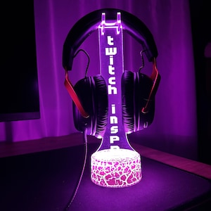 May include: A purple-lit headphone stand with the word "twitch inspa" vertically displayed. The stand holds a pair of black headphones with red accents. The base of the stand has a cracked pattern design. The background is a soft purple.