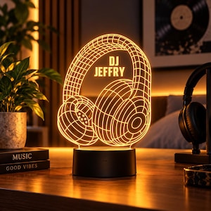 May include: An illuminated 3D acrylic lamp featuring a wireframe graphic of headphones with the text DJ JEFFRY centered in the design. The lamp is mounted on a solid black circular base and emits a warm orange glow.