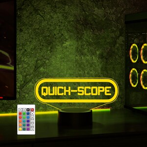 May include: A yellow neon sign with the text "QUICK-SCOPE" in a retro video game style. The sign is on a black base and has a remote control with multiple colored lights.