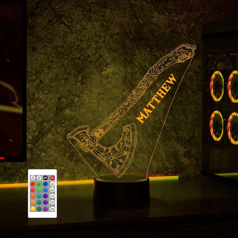 May include: A 3D printed night light shaped like an axe with intricate details and a personalized name "MATTHEW". The light is illuminated in a warm yellow color and has a remote control with multiple color options.