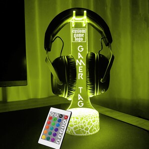 May include: A black and white gamer headset stand with a custom game logo and a remote control for changing the color of the LED lights. The stand is lit up in a bright yellow color.