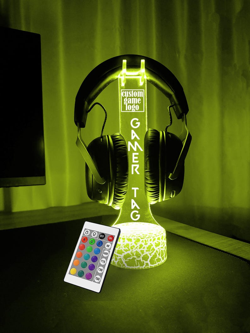 May include: A black and white gamer headset stand with a custom game logo and a remote control for changing the color of the LED lights. The stand is lit up in a bright yellow color.