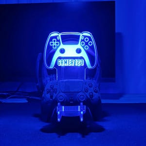 May include: A blue LED light shaped like a video game controller with the text "GAMER123" on it. The light is on a stand with a black video game controller.
