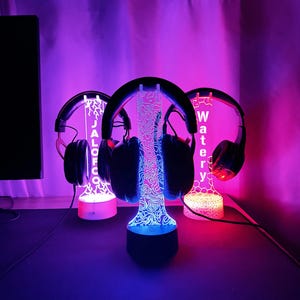 May include: Three illuminated headphone stands with headphones. Each stand has a unique design and text: "JALCO", "OFCO", and "WATERY". The stands are lit with blue, pink, and red lights, creating a vibrant display.
