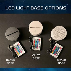 Op de afbeelding: Drie LED-lichtvoetjes met afstandsbedieningen. De voetjes zijn zwart, wit en wit met een gebarsten afwerking. Elk voetje heeft een USB-kabel en een afstandsbediening met meerdere kleur opties.