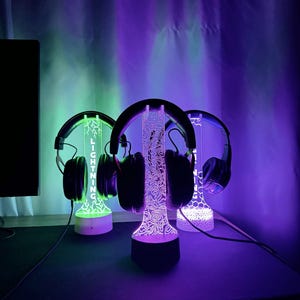 May include: Three illuminated headphone stands with headphones. The stands have a decorative design and the word "LIGHTNING" is visible on one. The stands are lit with green, purple, and white lights. The headphones are black.