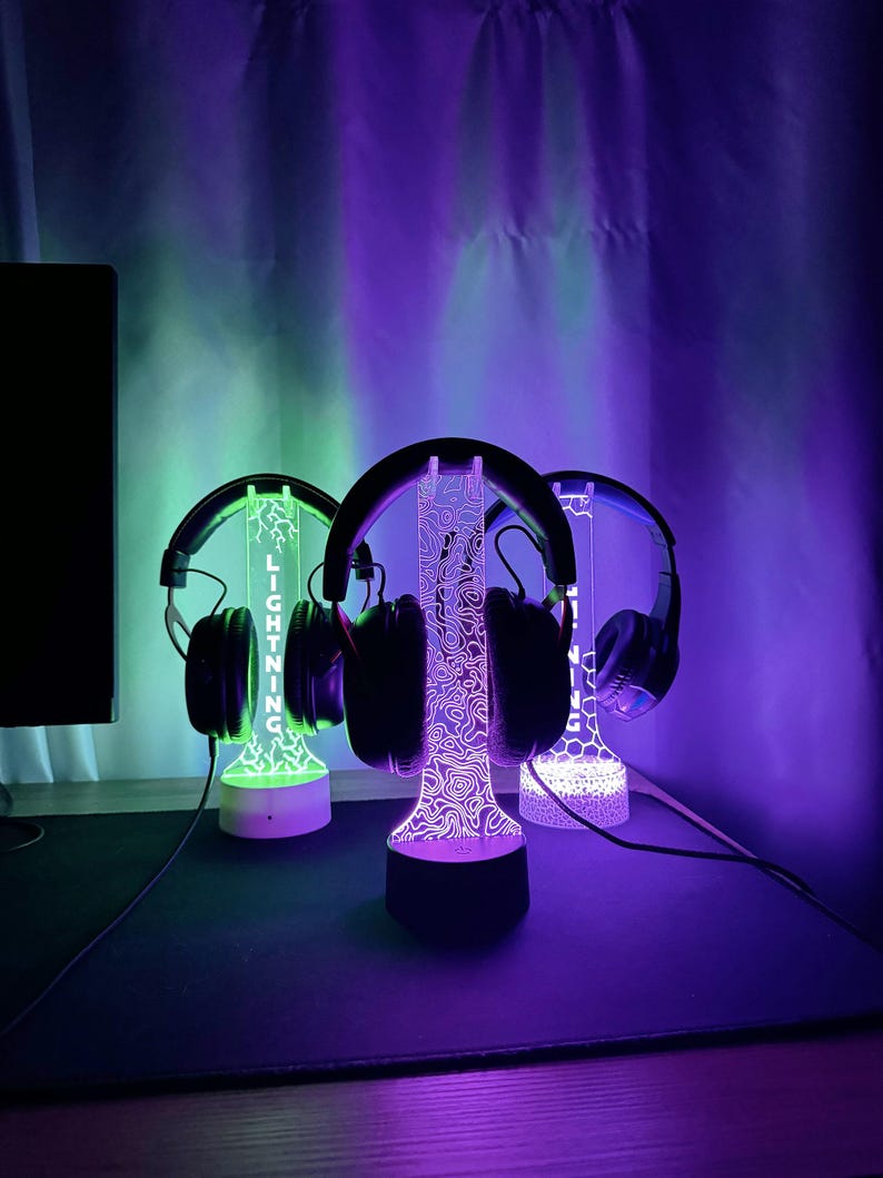 May include: Three illuminated headphone stands with headphones. The stands have a decorative design and the word "LIGHTNING" is visible on one. The stands are lit with green, purple, and white lights. The headphones are black.