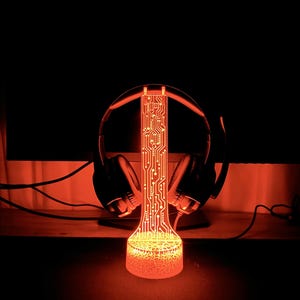 Op de afbeelding: Een rood LED-licht met een printplaatontwerp, gevormd als een standaard voor koptelefoons. Het licht is aan en verlicht de koptelefoons.