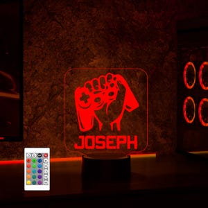May include: Red LED night light with a gamer hand holding a controller and the name "JOSEPH" underneath. The light is on a black base and comes with a remote control with multiple color options.